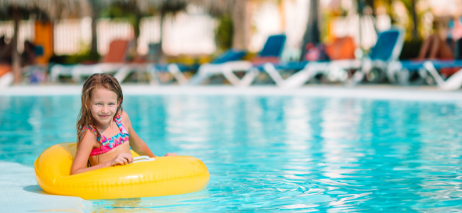 Niña flotando en una piscina con un inflable amarillo, con tumbonas y sombrillas en el fondo